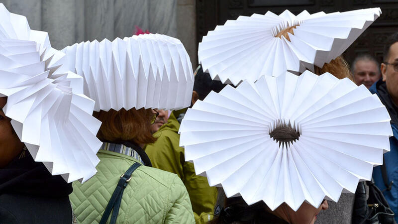 Four people wearing large, circular origami hats composed of accordion folds with a small hole at the center.