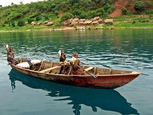 Three children in a row boat on water. In the background is the shore with trees, grass, and a village of thatch-roofed huts.