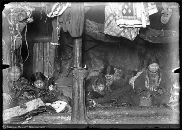 Five Koryak people seated in a wooden, interior space with a pole in the center and clothing hanging above.