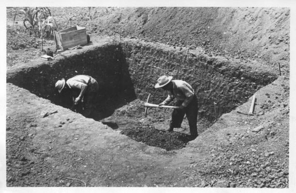 Two people wearing brimmed hats standing in a square excavation pit about six feet deep, working with double-headed pick axes.