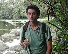A photo to the waist of man facing the camera wearing a tee shirt and back pack and holding perhaps a walking stick, with a swamp and trees in the background.