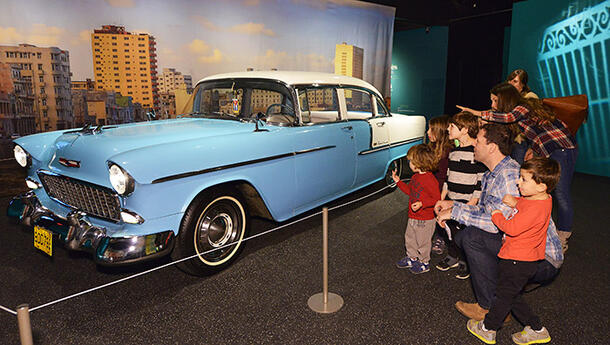 A group of adults and children look and point at a vintage '50s car parked in the Museum, placed against a photo backdrop of Havana.