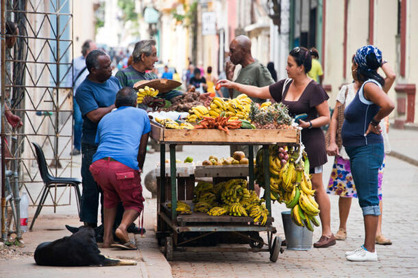 Customers crowd around a cart displaying bananas and other fruits and vegetables.