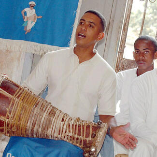 Man holds a batá drum and chants while two other worshippers stand nearby.