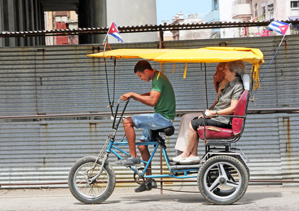 A young man pedals his bicitaxi and  transports two passengers.