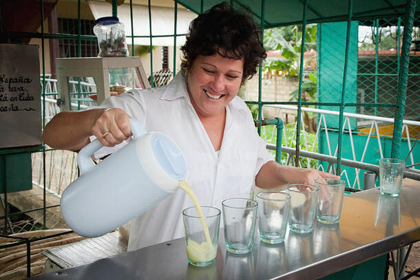 Woman uses a pitcher to pour fresh sugarcane juice into five glasses.