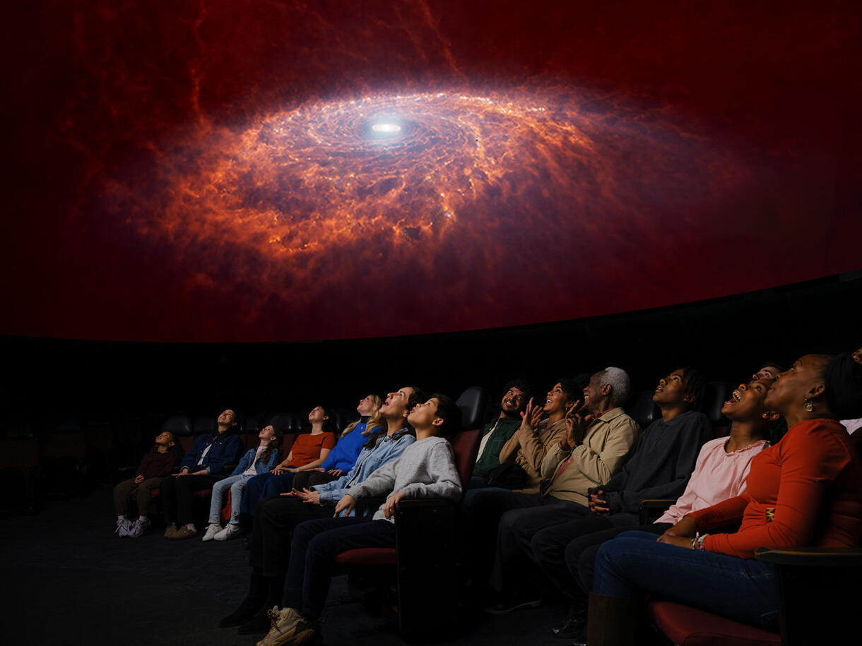 A seated audience, with about thirteen people visible, in the Hayden Planetarium look up in awe at a projection of a spiral galaxy.