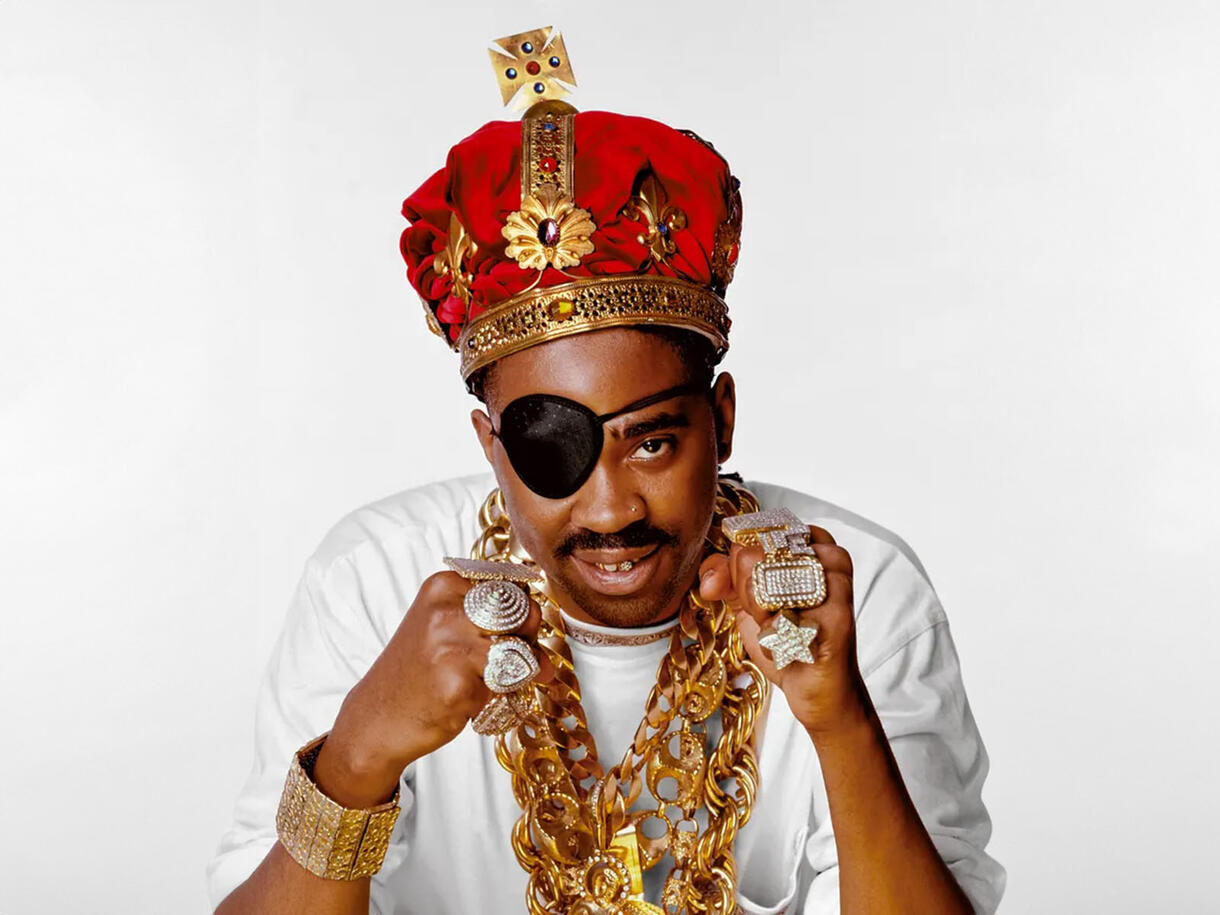 Slick Rick wearing an eyepatch over his right eye, a large crown, heavy gold necklaces, and over 7 large rings.