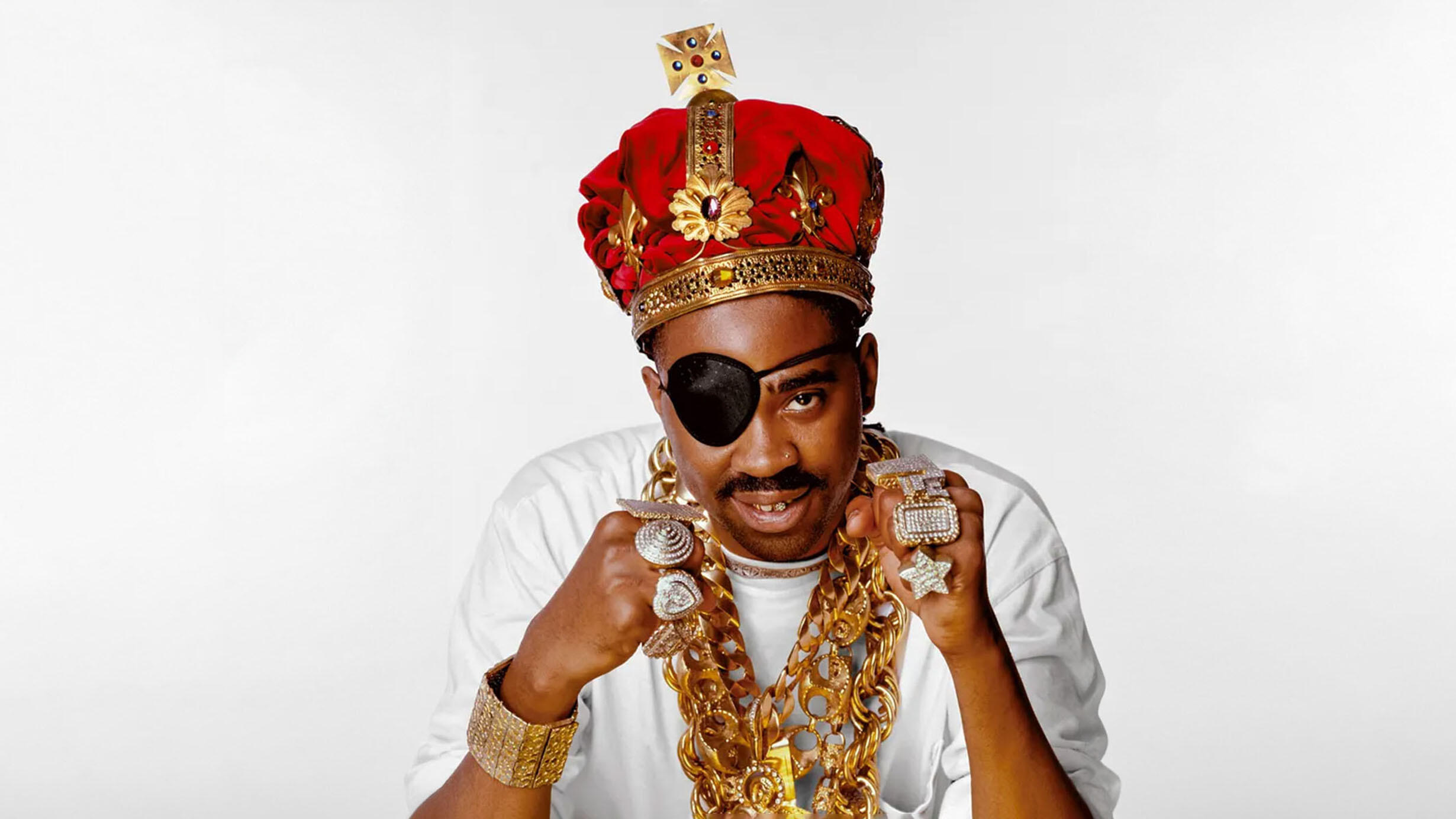 Slick Rick wearing an eyepatch over his right eye, a large crown, heavy gold necklaces, and over 7 large rings.