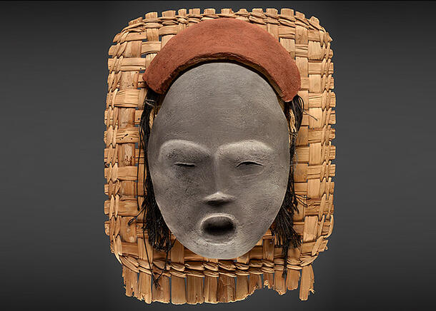 Clay mask with simply executed closed eyes, slim nose and open mouth rests atop a straw mat.