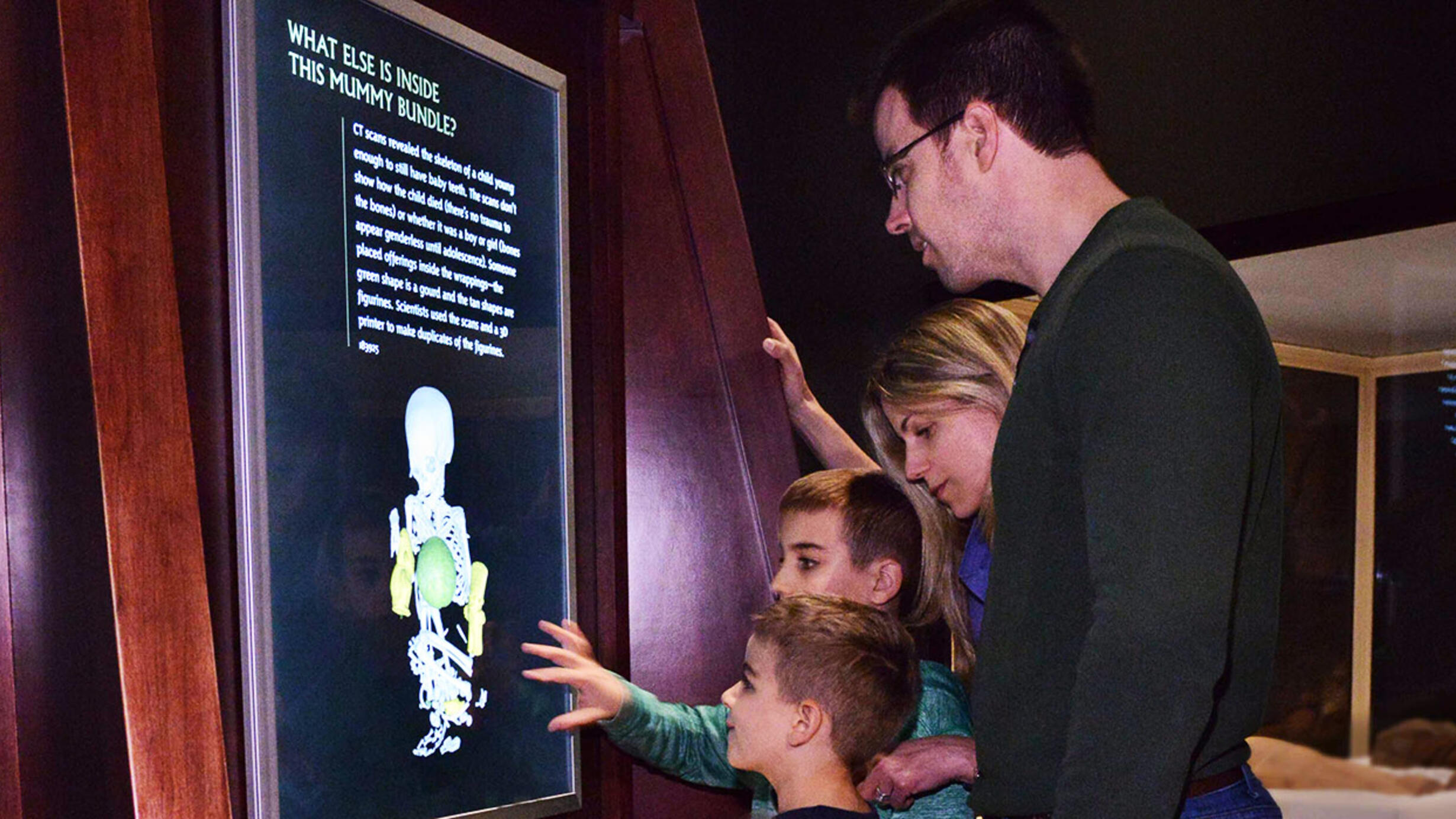 Two children and two adults gather around a digital touch screen placed on the wall that shows views from a CT scan of a peruvian mummy.