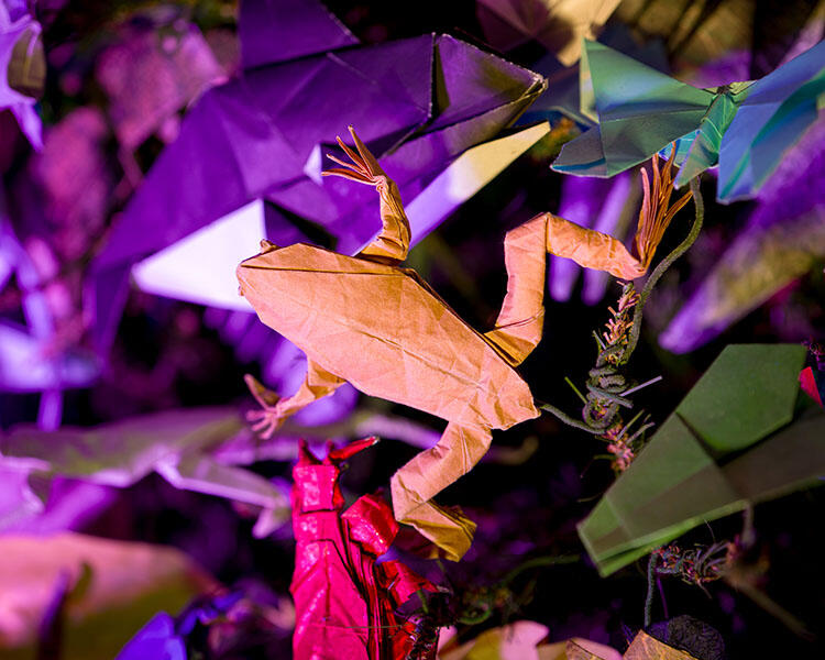 A frog ornament made with origami (folded paper) perched on an artificial evergreen tree, surrounded by other paper ornaments.