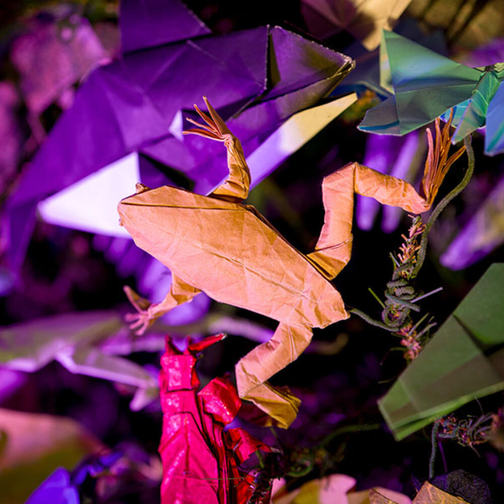 A frog ornament made with origami (folded paper) perched on an artificial evergreen tree, surrounded by other paper ornaments.