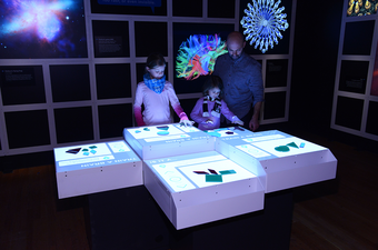 Children and adults gather around four lit tabletop surfaces that hold that plastic shapes.