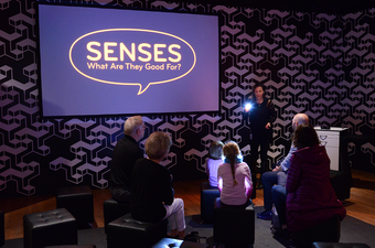 A small audience sits in front of a presenter and a screen.