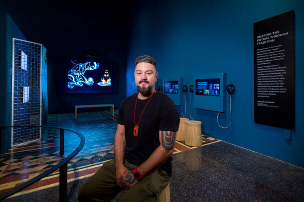 Artist Bracken Hanuse Corlett sitting on a stool in the Shaping the Future Through Traditions exhibit. 