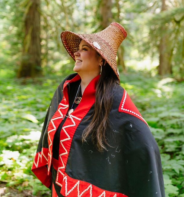 Artist Danika Saunders wearing a traditional outfit and woven hat, standing outside in a forest. 