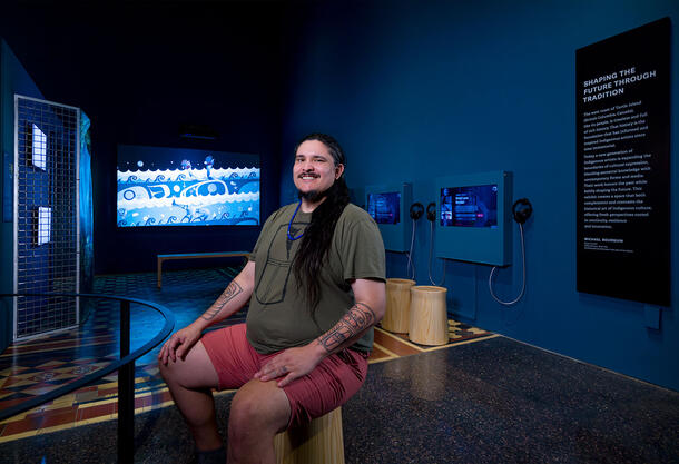 Artist Dustin McGladrey sitting on a stool in the Shaping the Future Through Traditions exhibit. 