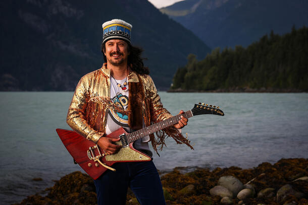 Artist Wil Uks Batsga Galaaw (Jeremy Pahl) wearing a traditional headpiece, shiny jacket, and holding an electric guitar, standing outside on the rocky shores of a lake.