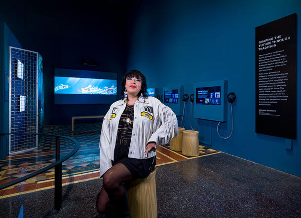 Artist Kristi Lane Sinclair, wearing a fringed jacket, sitting on a stool in the Shaping the Future Through Traditions exhibit. 