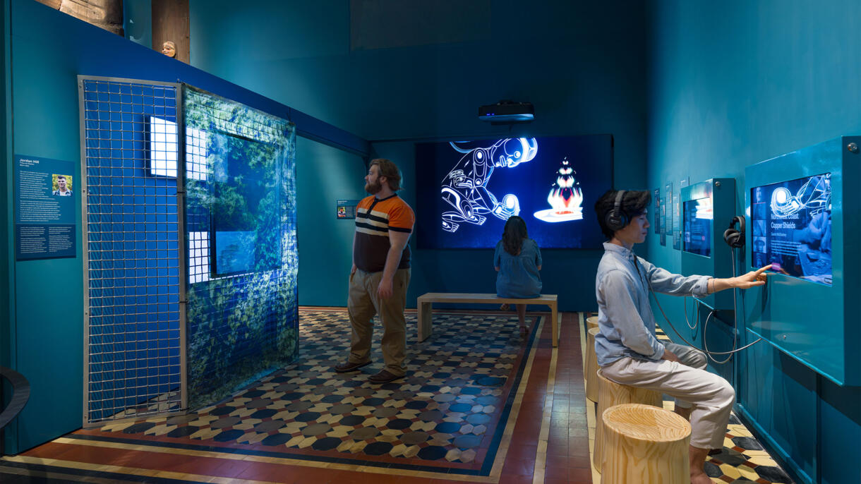 Three Museum visitors interact with video and audio components of the exhibitShaping the Future Through Tradition in the Northwest Coast Hall.