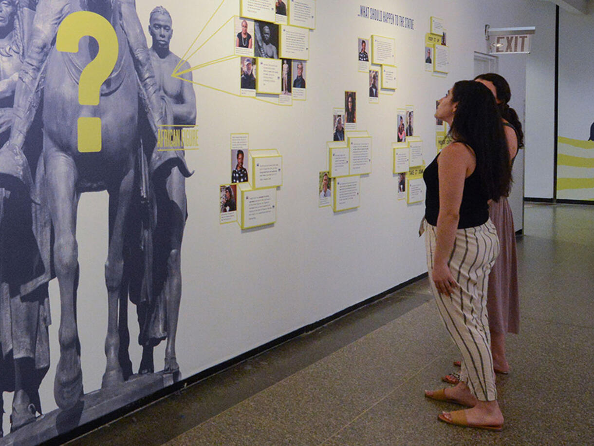 People standing in a indoor exhibition room, looking at images on a wall.