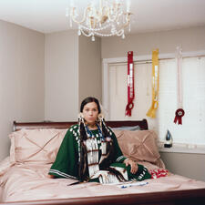 Danielle Fin sits on a bed wearing traditional dress, her pageant ribbons hang from the wall behind her.