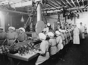 A dozen woman are seated at an assembly line in a factory, with two supervisors standing behind them.