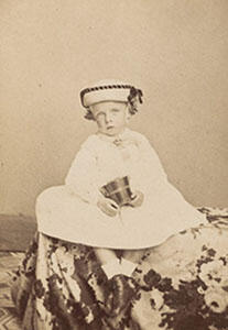 A toddler-aged Theodore Roosevelt sits for a formal portrait wearing a sailor-style hat and holding a small bucket.