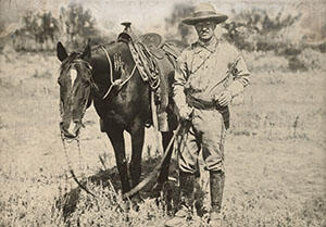 Theodore Roosevelt stands next to a horse in a field.