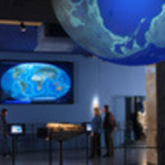 A museum hall with a massive model of the Earth suspended from the ceiling, and a large electronic screen on the wall showing a map of Earth.