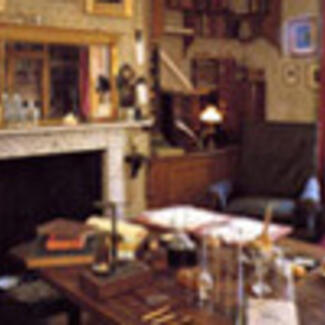 A drawing room with a fireplace and a large table covered with books and other objects.