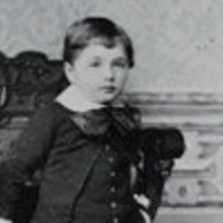 An old black and white photo of young boy in formal attire, with a buttoned-down coat, white collared shirt, and a bow tie.