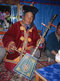 An older man in traditional clothes kneels on the floor with a stringed instrument and bow.