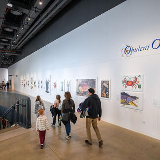 Museum visitors enter an open walkway that has a railing on the left and walls on the right that display large vintage illustrations of ocean life.