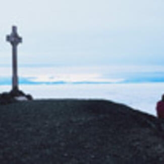 A Cross monument on a hill with a large body of water in the background, and a person seated on the ground.