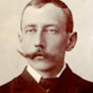 A black and white photo of a man with short hair, in a formal jacket and necktie, with a clean-shaven face but for a large handlebar mustache.