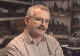 A man with eyeglasses and a mustache seated indoors posing for the camera.