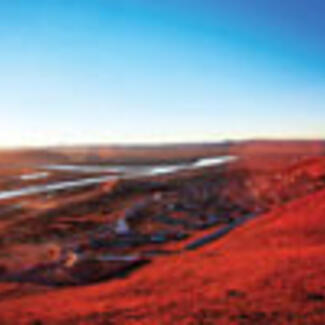 A wide shot of the Sichuan Basin, China. The rolling fold-and-valley terrain appears red, and has a wide clear blue sky above it and a river flowing through it in the distance.