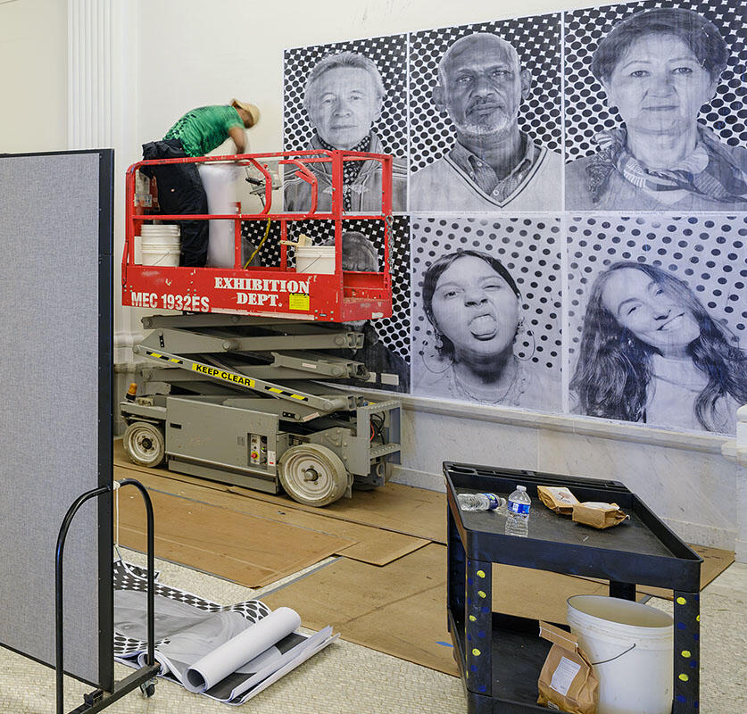 Exhibitions team member stands on a lift to hang larger-than-life photo portraits on the walls lining the Ellen Futter Gallery.