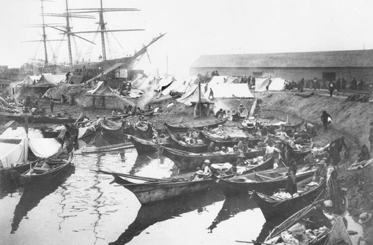 A wooden ship and many smaller wooden dinghies, many full of people, docked at a lively port where tents are pitched by the ship.