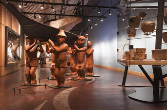 Larger-than-life, terracotta colored sculpture of 6 people carrying a canoe in a museum gallery space, with glass case full of baskets to the right.