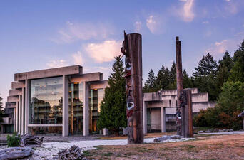 Modern building with T-shaped beams over glass window among trees and bright sky with four carved, wooden poles outside.