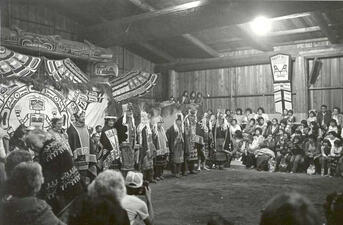 Men and women in elaborate robes and headdresses stand in front of large bird figures and patterned backdrop before a large audience in a wooden hall.