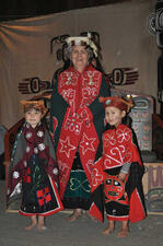 Older woman in headdress and elaborate dress decorated with beaded stars and butterflies stands between two kids in similar dress and robes.