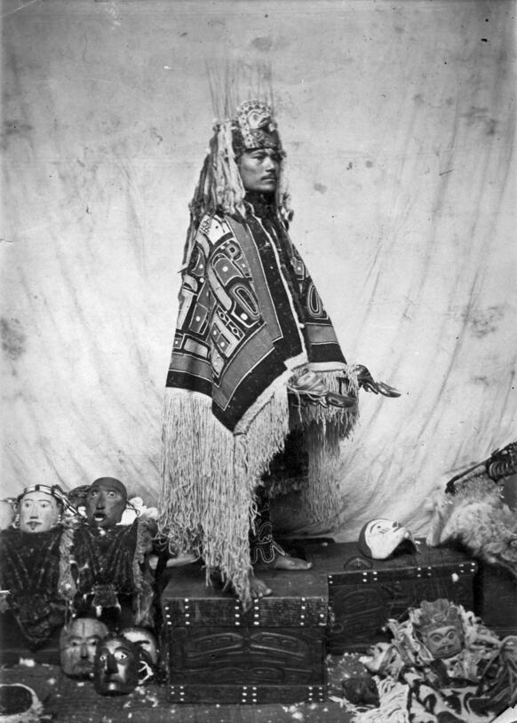 Man in elaborate headpiece and fringed, patterned robe stands on carved chest holding carved rattle against plain background, with masks on the floor.