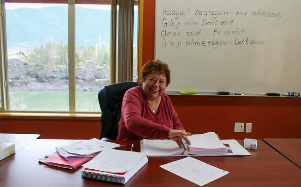 Teacher seated at long table, in front of window and whiteboard displaying translated phrases, with open binder and papers on table in front of them.