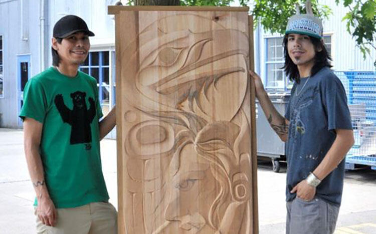 Nunuusxli (Chazz Mack) and Wiiqa7ay (Lyle Mack) stand outside holding a carved wooden door in between them.