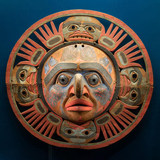 Carved mask depicting face at center with curved nose surrounded by four additional faces and hand-shaped carvings radiating out in circle.