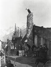 Historical image of 25 adults, children and 1 dog sit and stand in front of wooden A-frame building with carved pole in front featuring bird on top.